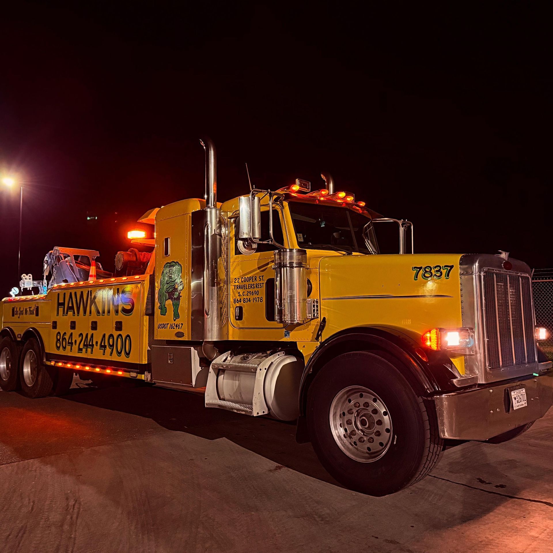 Yellow tow truck with flashing lights parked at night on a street, towing a car.