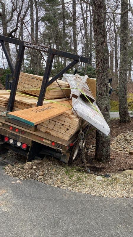 Overloaded flatbed trailer tipped into roadside trees, with lumber and debris spilling off.