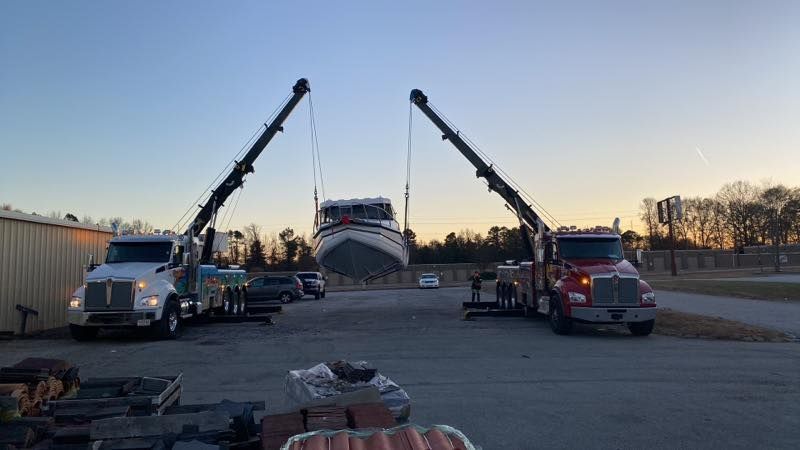 Two tow trucks lifting a boat at dusk in a parking lot