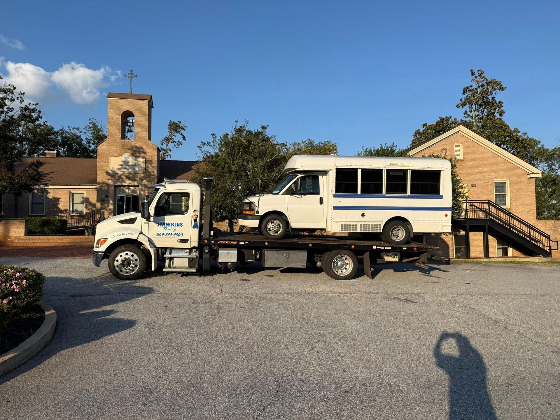 White utility truck carrying a beige van on a flatbed in front of a brick building under a blue sky