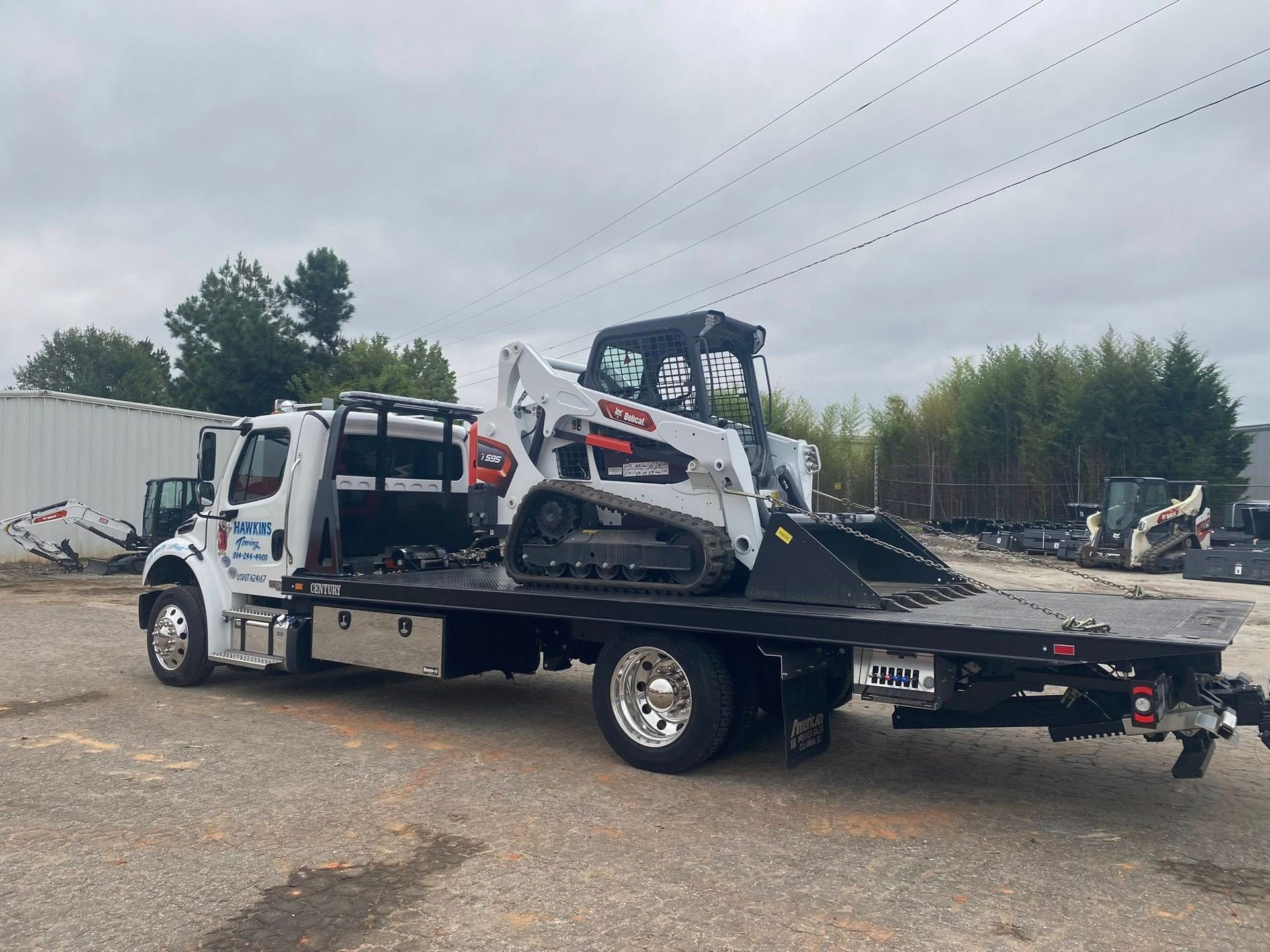 Tow truck carrying a white Bobcat skid steer on a flatbed in a gravel lot
