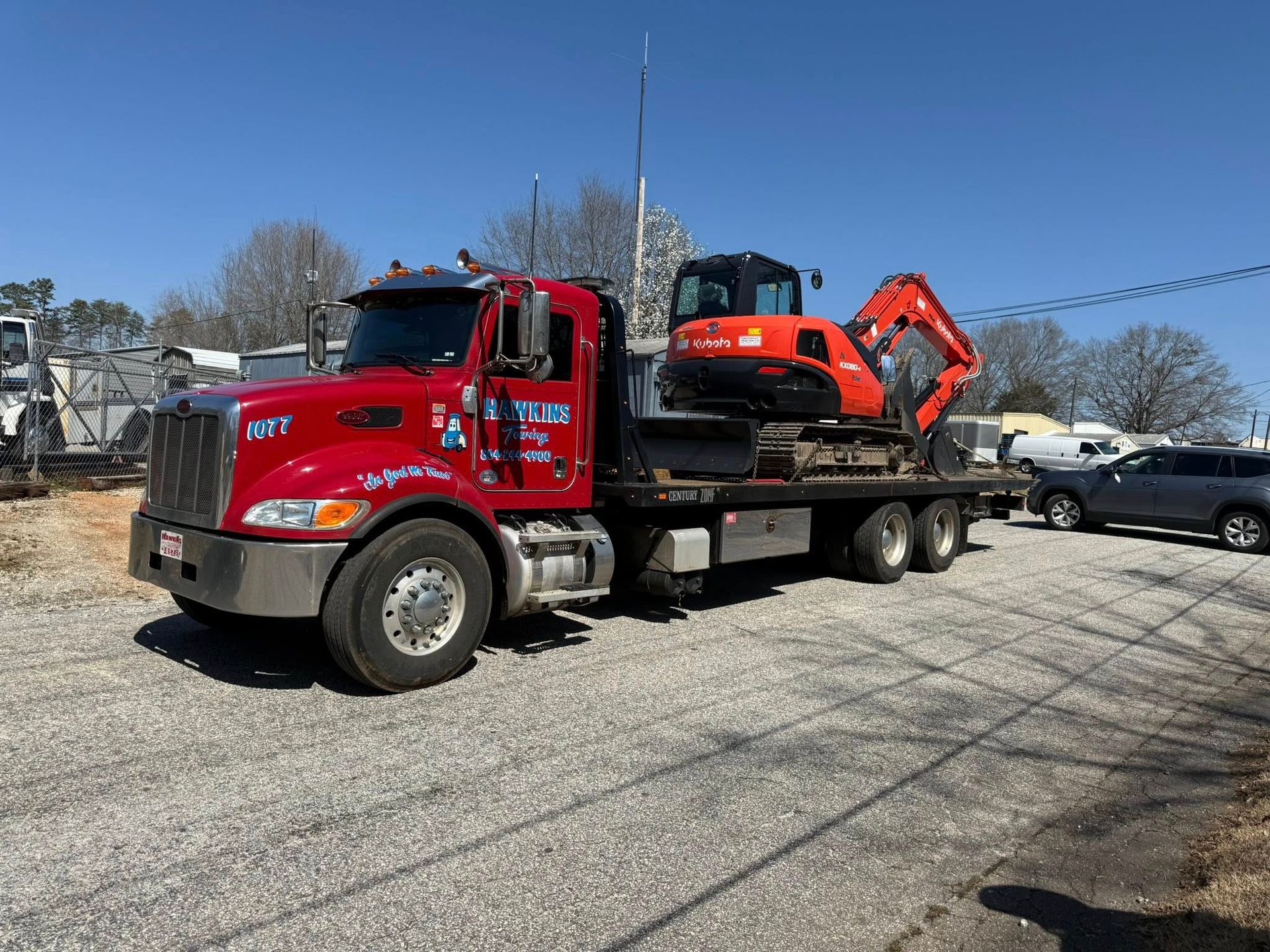 Red flatbed truck hauling an orange excavator in a gravel lot on a sunny day
