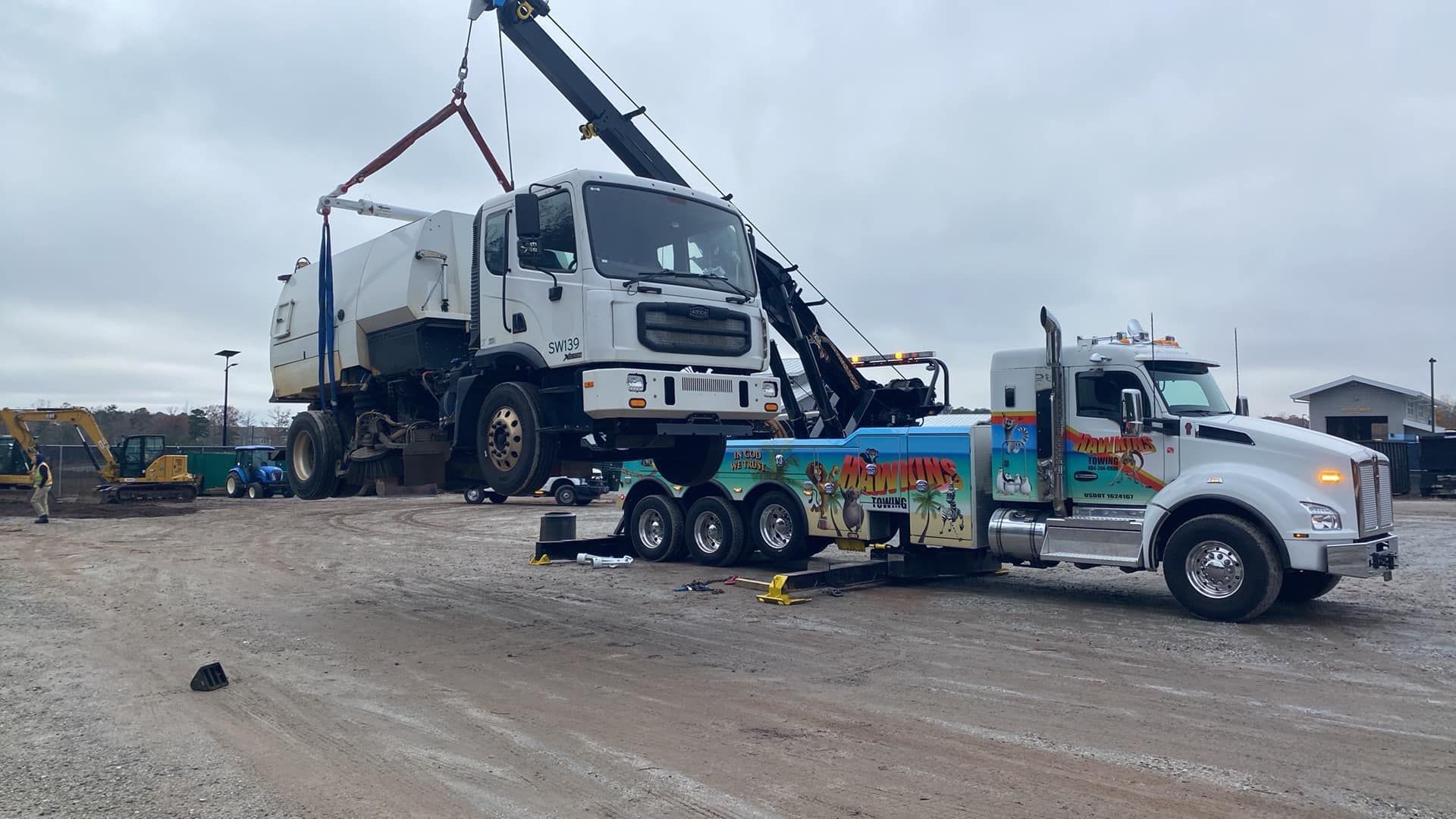 Tow truck lifting a white garbage truck beside a blue truck on a muddy roadside.