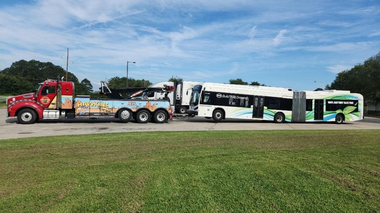 Red tow truck hauling a long articulated bus on a grassy roadside under a blue sky
