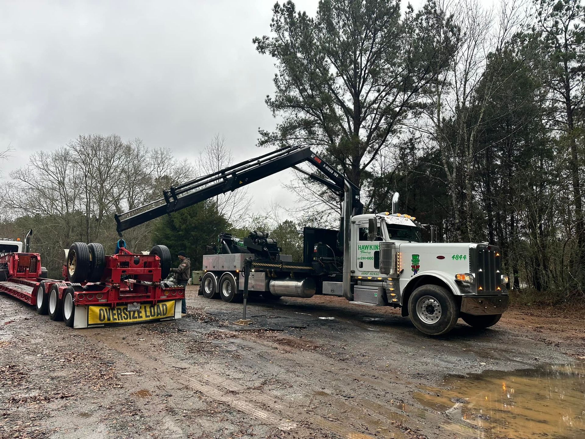Tow truck lifting a white semi-truck cab in a muddy lot under overcast skies