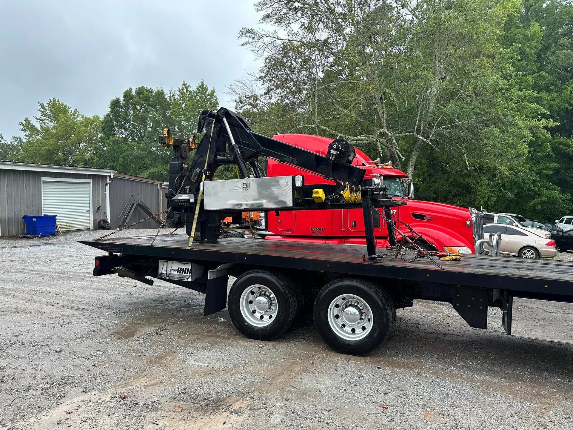 Red tow truck with crane on a flatbed trailer in a gravel lot beside trees and a garage.