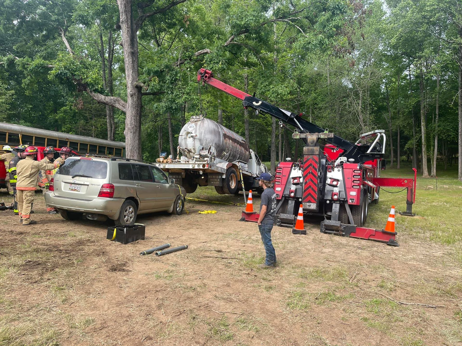 Tow truck lifting a silver SUV in a wooded area, with firefighters and cones nearby.