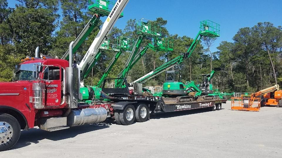 Red semi hauling green boom lifts on trailers in a lot beside orange construction equipment