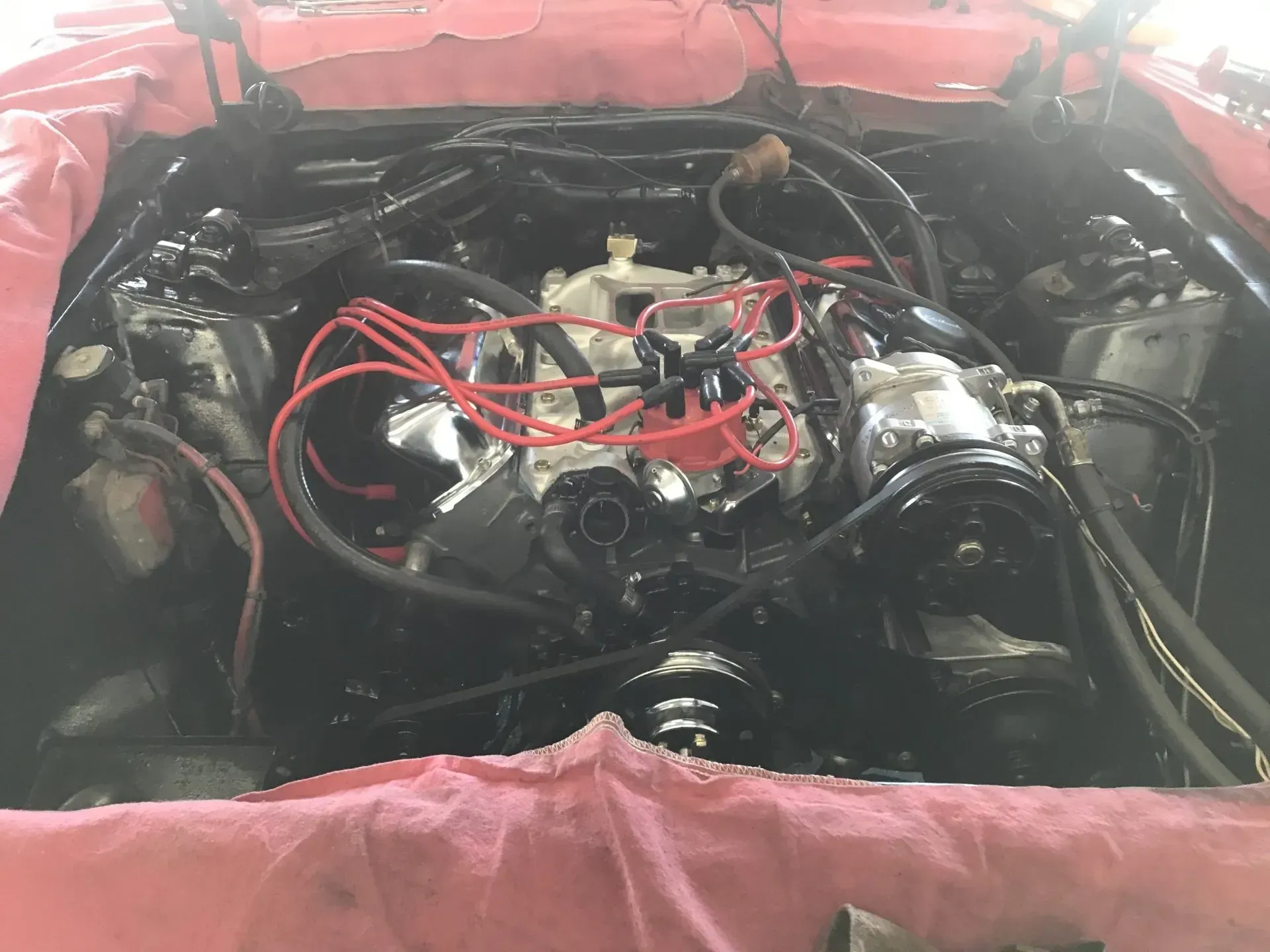 Car engine bay with red wiring and chrome components, viewed from above with a pink fender cover around it
