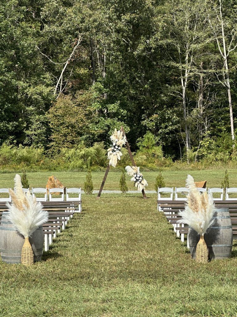 Outdoor wedding aisle with white chairs, decorative pampas grass, and forest backdrop