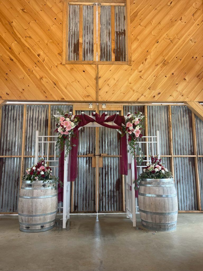 Rustic barn entrance with floral arch, burgundy drapes, and two wooden barrels holding flowers
