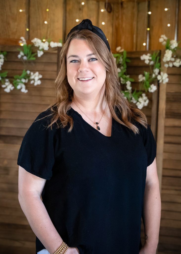 Woman in a black top smiling in front of a wooden wall with white floral decorations.