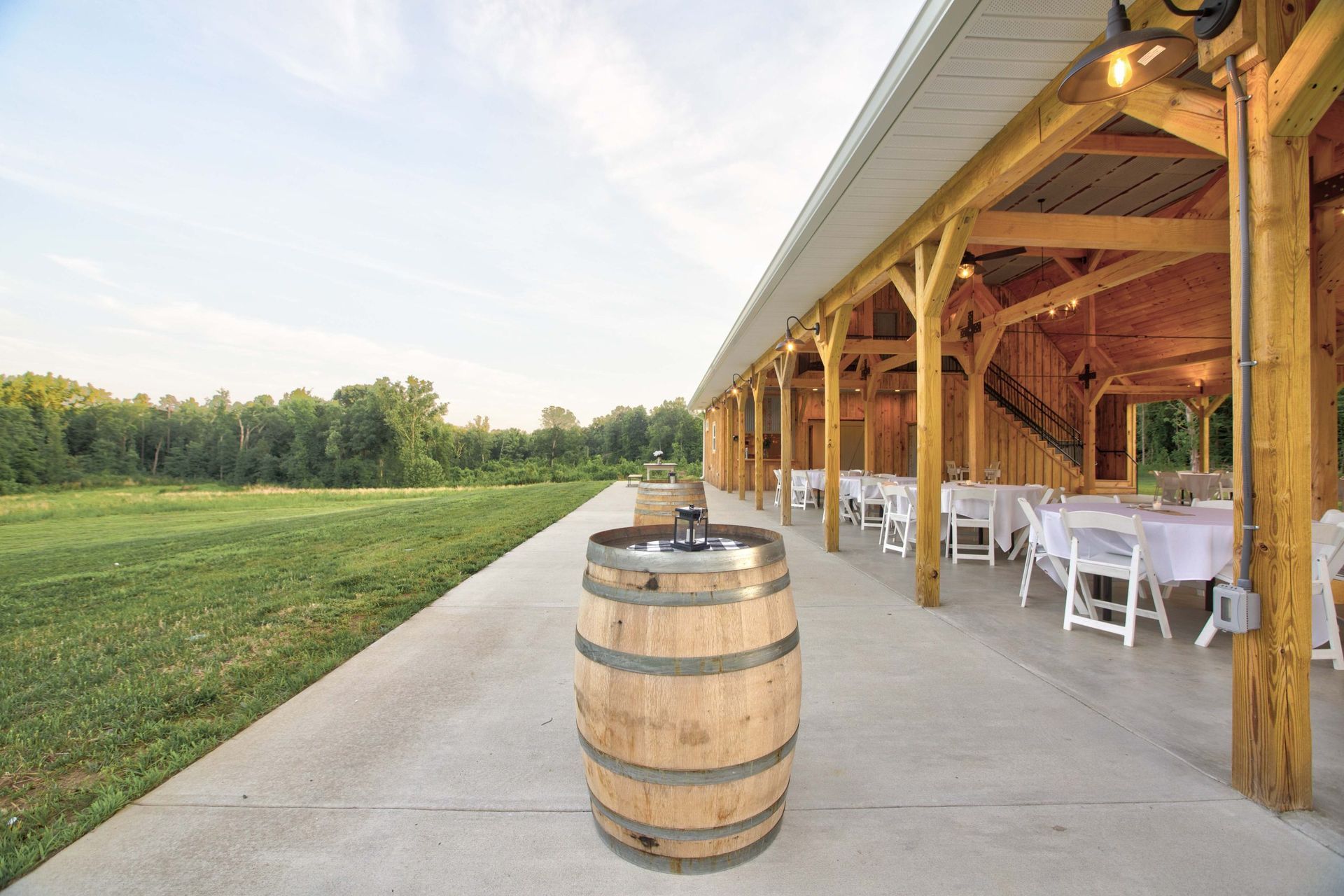Covered outdoor pavilion with white chairs beside a wooden barrel on a concrete patio overlooking a grassy field