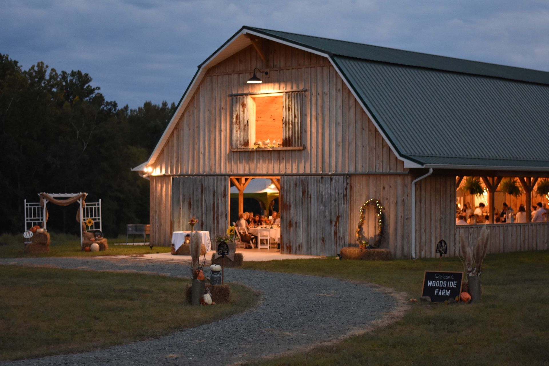 A lit rustic barn at dusk with a gravel path, outdoor decorations, and warm lights glowing inside.