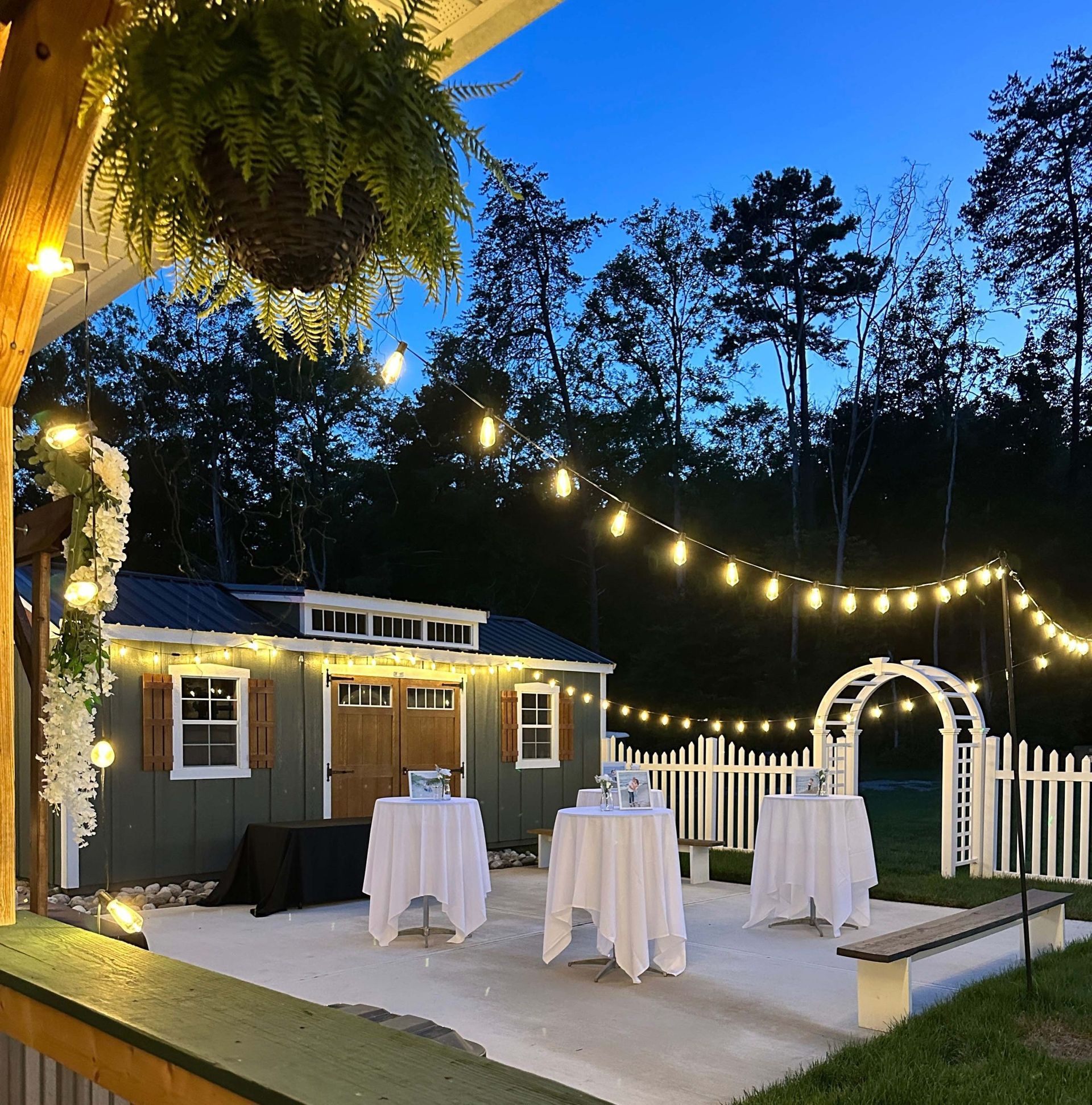 String-lit outdoor patio with cocktail tables, white arch, and a small garden shed at dusk