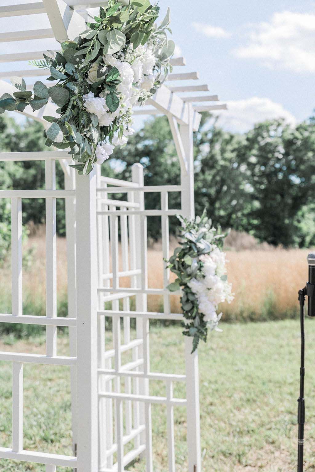 White floral wedding arch with greenery and hanging blooms outdoors in a garden.