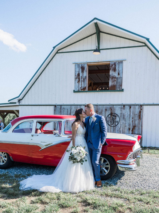 Bride and groom kissing beside a red vintage car in front of a white barn