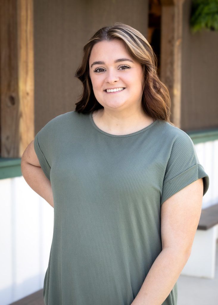Woman standing outdoors in a green top, smiling in front of a wooden porch.