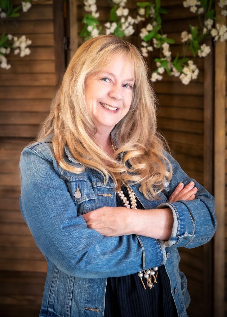 Smiling woman in a denim jacket with arms crossed, standing before wood paneling and flowers