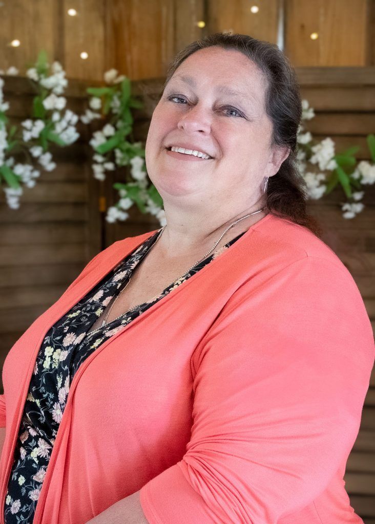 Smiling woman in a coral cardigan standing indoors before wood paneling and white floral decor