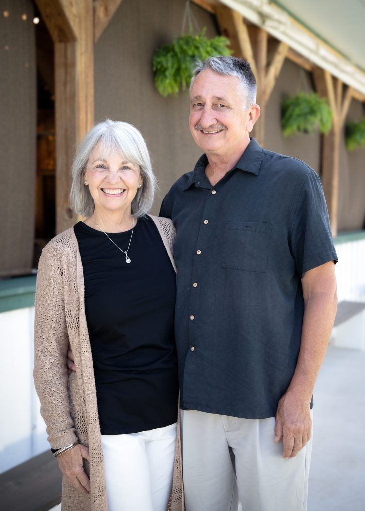 Smiling couple standing outdoors in front of a wooden building with hanging green plants.