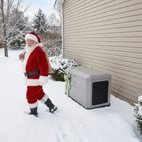 Santa standing in snow beside a house and outdoor unit, holding a shovel