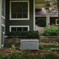 Stone retaining wall with a gray utility box in front of a house with dark siding and windows