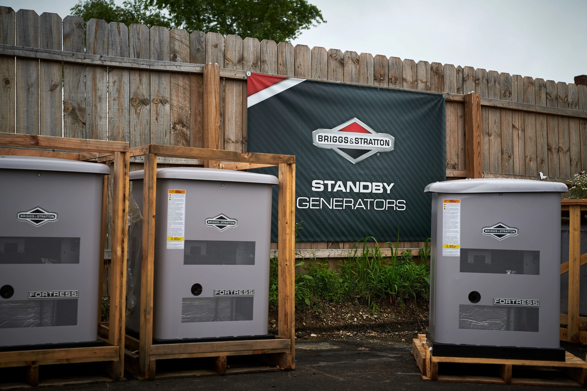 Three gray standby generators lined up outdoors in front of a wooden fence and banner.