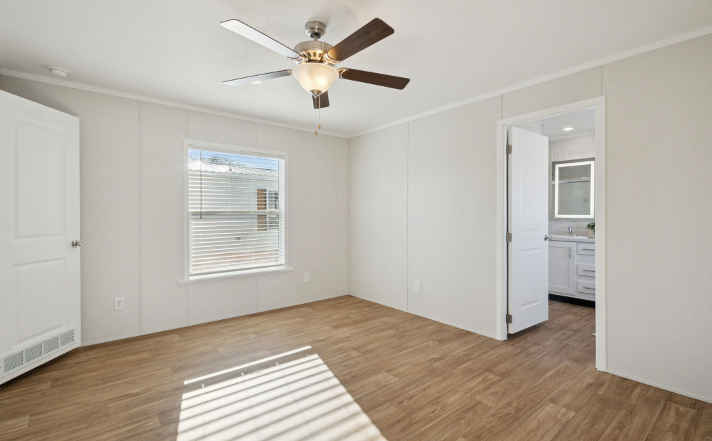 Empty bedroom with white walls, wood floor, ceiling fan, window blinds, and an open doorway to a bathroom.