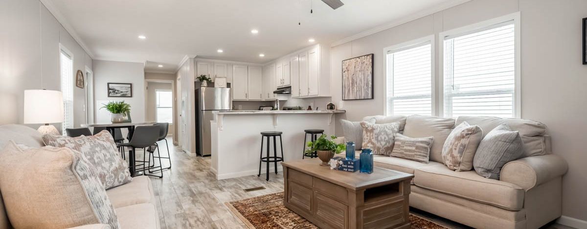 Living room with a kitchen in the background. White sofas and kitchen cabinets. Wooden coffee table with decorations.