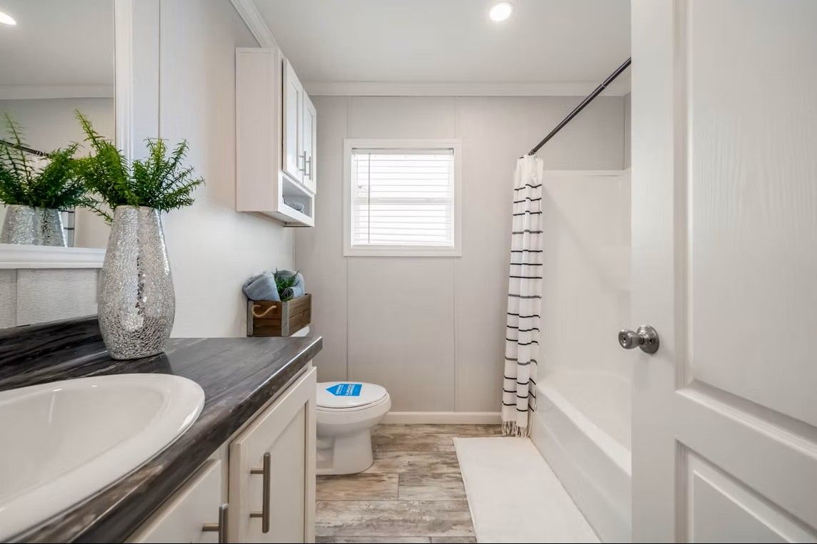 White bathroom with a sink, toilet, and bathtub. The room has a window, cabinet, and a shower curtain.