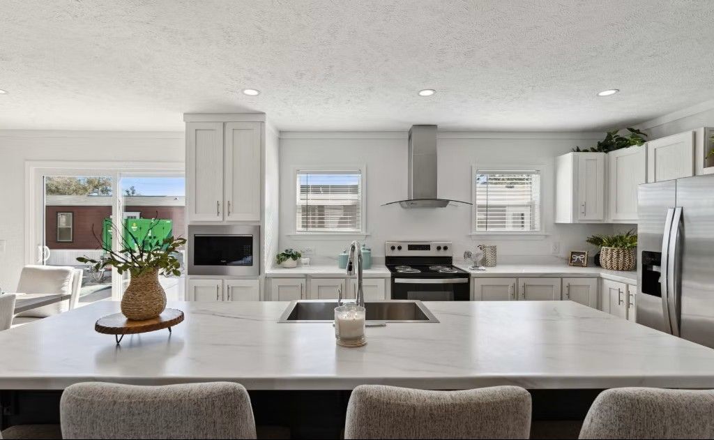 Modern white kitchen with large island, stainless steel appliances, and large windows.