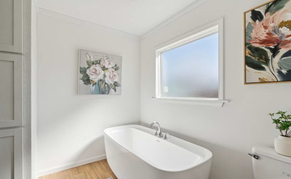 A white bathroom with a freestanding tub, window, and floral art on the wall.