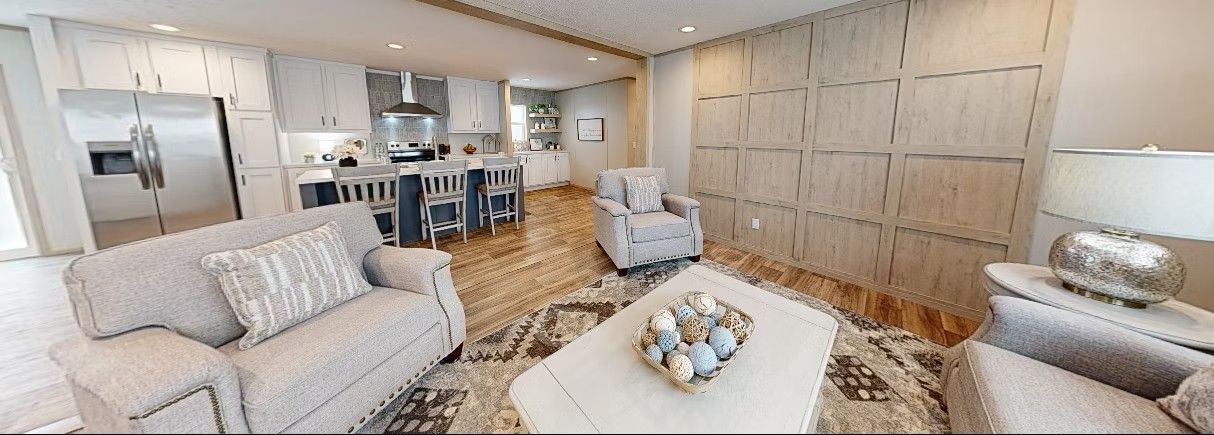 Living room with light-colored sofa, chair, and coffee table. Kitchen with bar in the background. Light wood floors.
