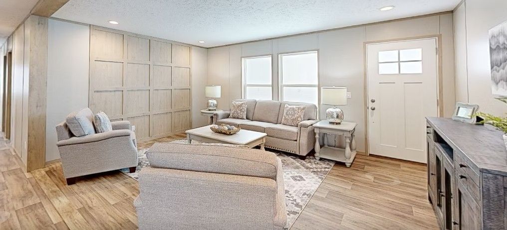 Living room with light-colored furnishings: sofa, armchair, ottoman, coffee table, and side tables. Wooden floor and panel wall.