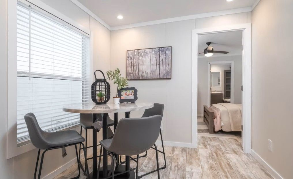 Dining area with round table, gray chairs, window with blinds, and doorway to a bedroom.