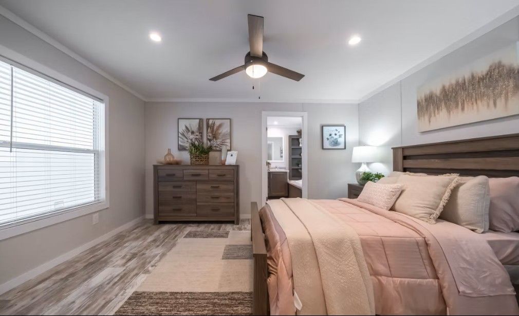 Bedroom with bed, dresser, art, window, and ceiling fan. Pink bedding, gray walls, light wood furniture.