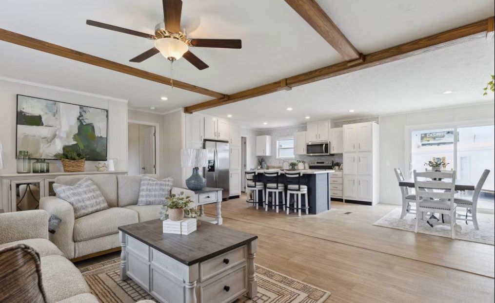 Open-concept living space with a kitchen, living area, and dining space; light wood floors, beige furniture, and brown ceiling beams.