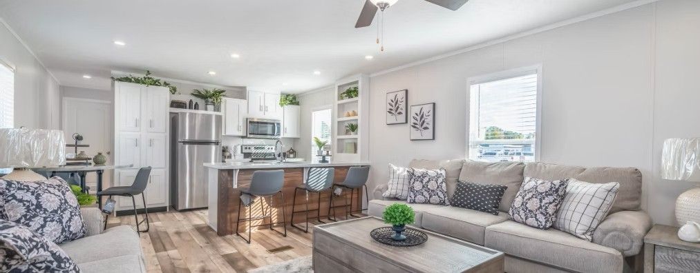 Interior of a modern living room and kitchen; white walls, wooden floors, gray furniture, and island with barstools.