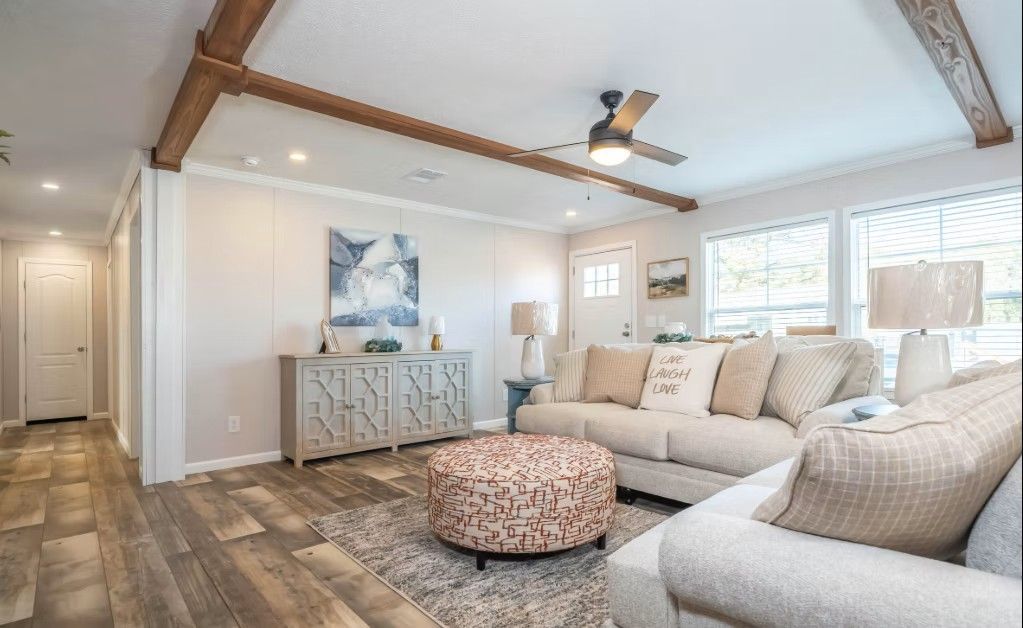 Bright living room with white walls, wood beams, a light-colored sofa, and an ottoman on a patterned rug.