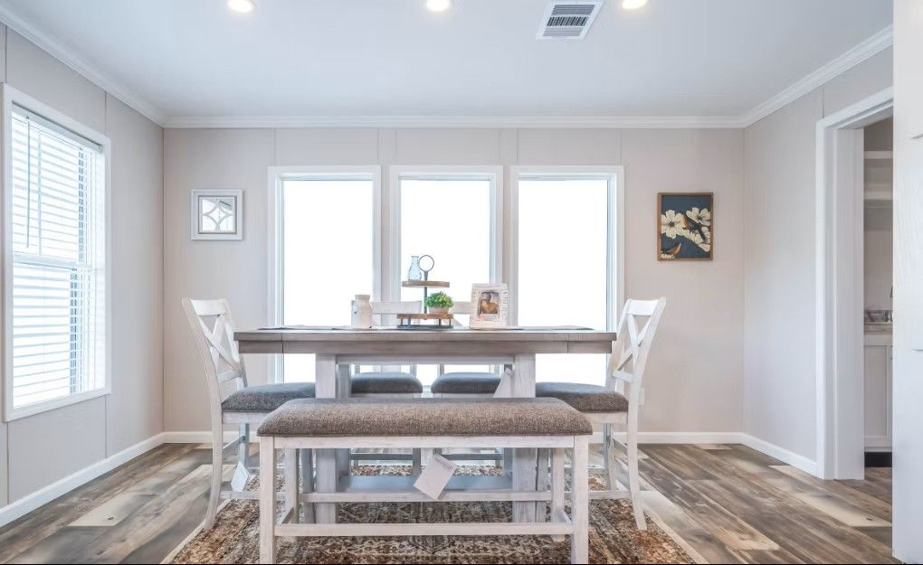 Dining area with wooden table, chairs, bench. Windows, artwork, and light-colored walls.