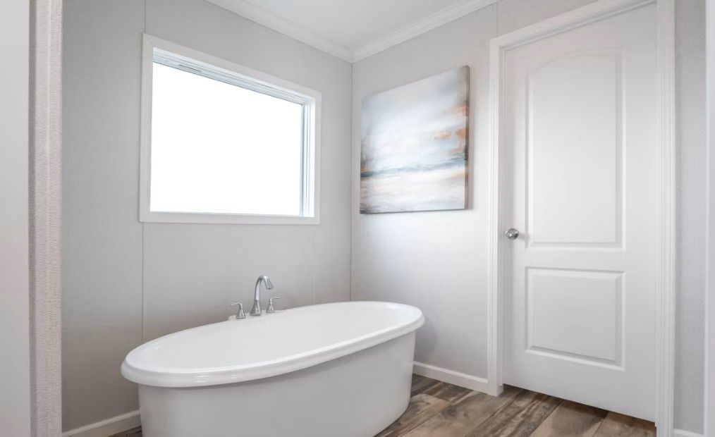 Bathroom with a white oval bathtub, window, artwork, and white door. Gray walls and wood-look flooring.