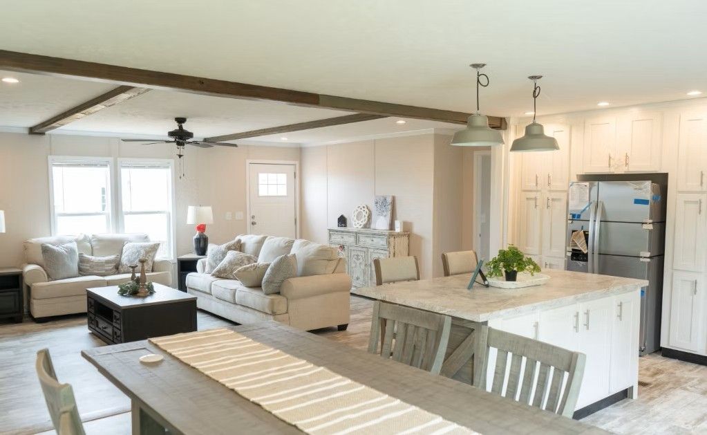 Open-concept living space with a kitchen island, dining table, and two couches. White walls, wooden beams, and neutral furniture.