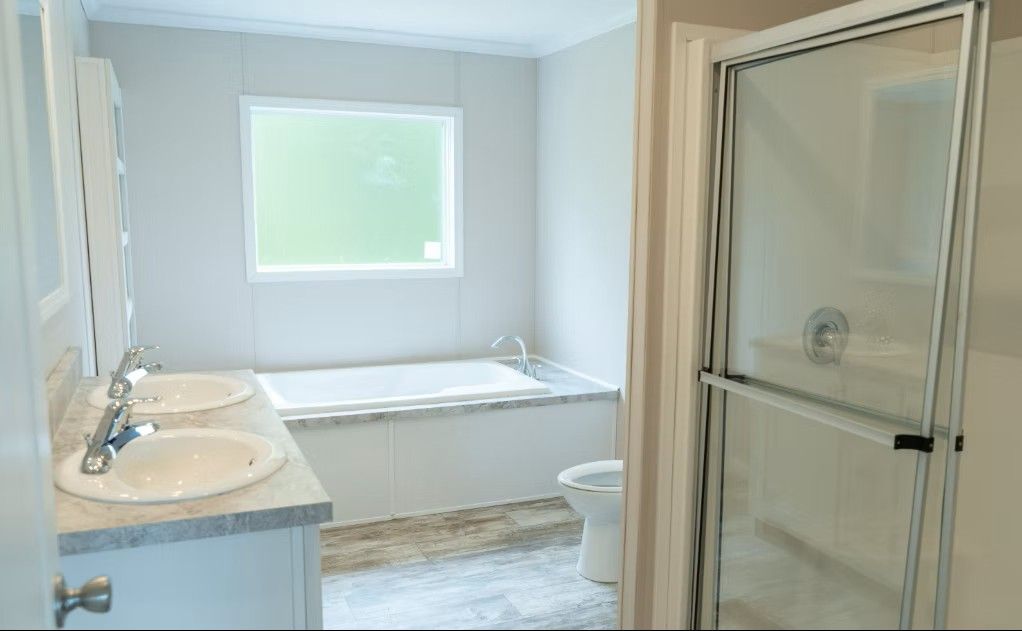 Bathroom with double sinks, bathtub, shower, and toilet. Light-colored walls and floor. Window in background.