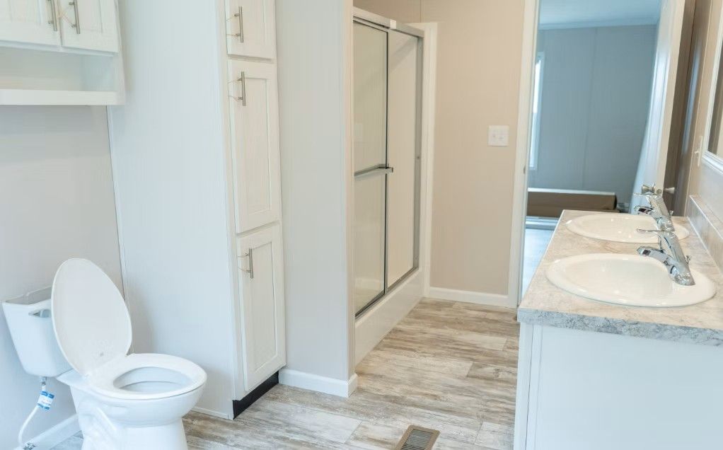 Bathroom with a toilet, vanity with two sinks, shower, and storage cabinet; light colors.