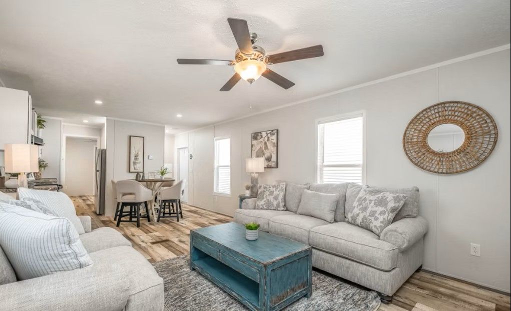 Spacious living room with light-colored couches, blue coffee table, dining area, and decorative ceiling fan.