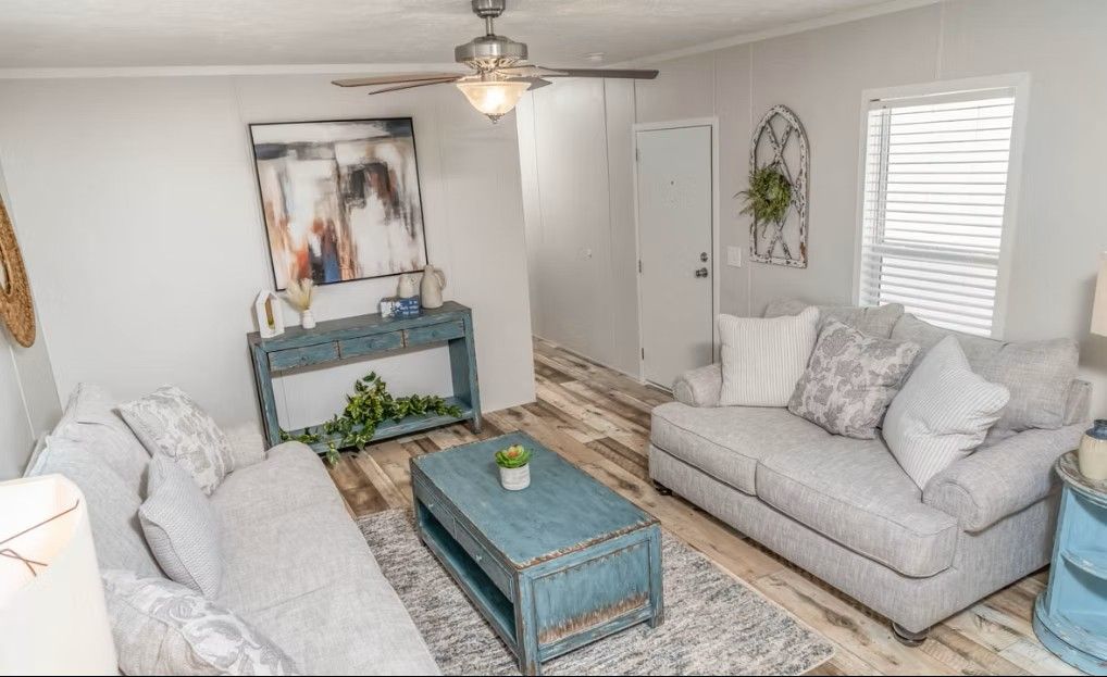 Living room with light-colored walls, gray couches, distressed blue furniture, and a rug on wood-look flooring.