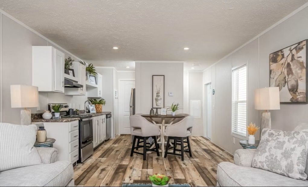 Interior view of a small living space with a kitchen, dining area, and seating. Wooden floor, white walls and cabinets.