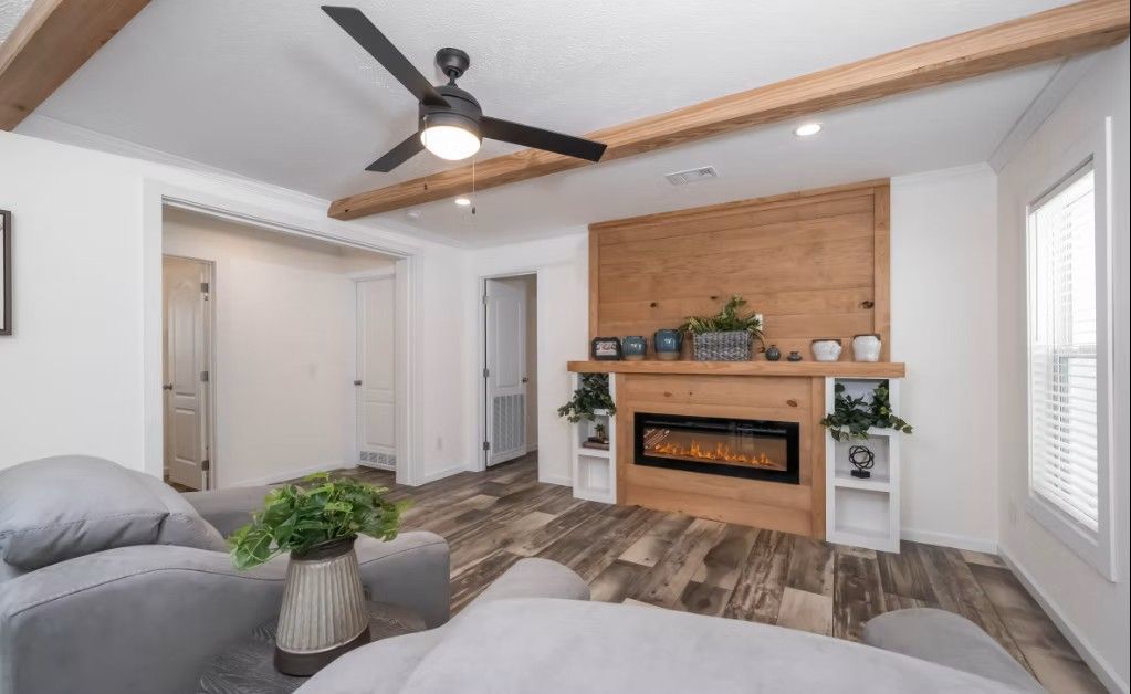 Living room with fireplace, wood accent wall, grey sofa, wooden beams, ceiling fan, and hardwood floors.