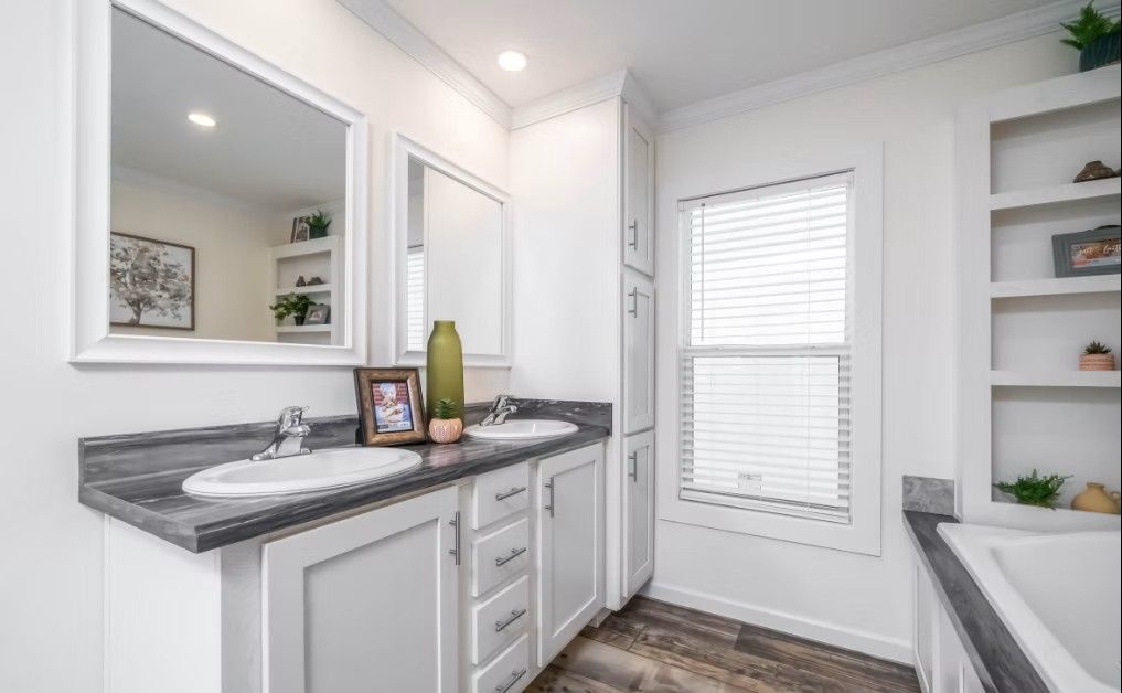Bathroom with double vanity, white cabinets, grey countertop, mirror, and window with blinds.